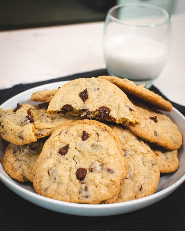 one of the best sweet treats, chocolate-chip cookies placed in a white plate with half glass of milk in the background